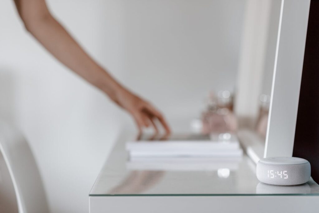 A smart speaker with a digital clock display placed on a minimalist desk setting, hand in background.